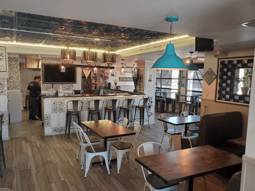 A modern restaurant interior with wooden tables, white chairs, a bar area, and decorative tiles.