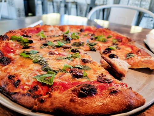 A close-up of a pizza topped with cheese, olives, and fresh basil, with a slice missing on a metal tray.