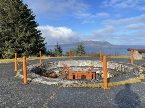 A circular stone structure with a rusted metal feature, surrounded by trees and overlooking a body of water and mountains.