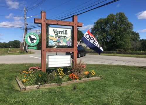Sign for Verrill Farm with "OPEN" flag, surrounded by flowers and greenery, on a sunny day.