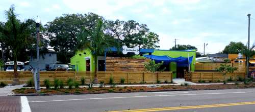 A colorful dog-themed restaurant with outdoor seating, surrounded by greenery and wooden fencing.
