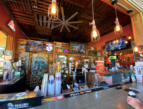 A vibrant bar interior with colorful decor, multiple TVs, and a variety of drinks displayed on the counter.