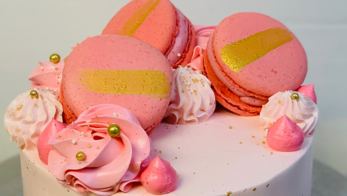 A pink cake topped with macarons, gold accents, and decorative swirls of frosting.