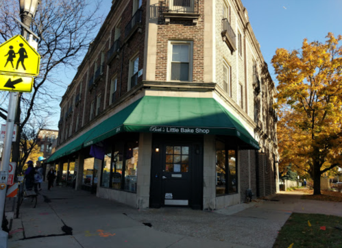 A corner bakery with a green awning, surrounded by trees and a sidewalk, in a brick building on a sunny day.