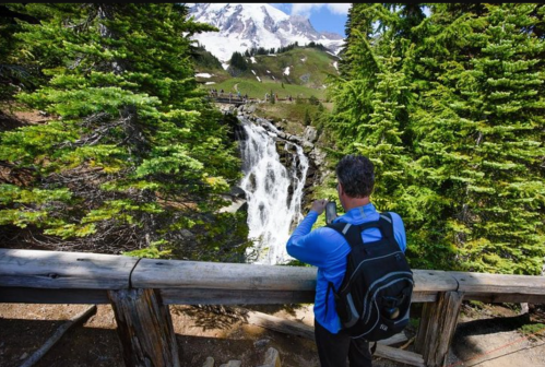 A person in a blue shirt stands by a wooden railing, taking a photo of a waterfall surrounded by lush greenery.