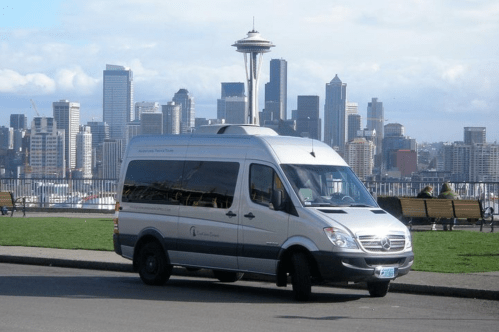 A silver van parked in front of a city skyline featuring tall buildings and a prominent space needle.