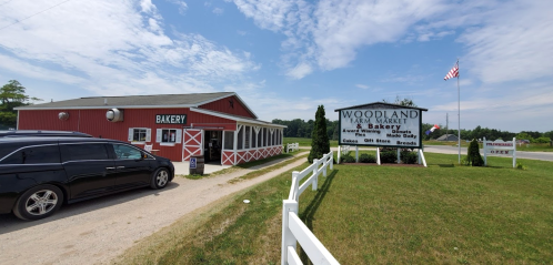A red barn-style bakery with a sign for Woodland Farm Market, featuring a grassy area and a clear blue sky.