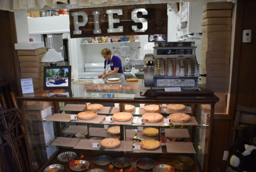 A display case filled with pies, with a vintage cash register in front and a baker working in the background.