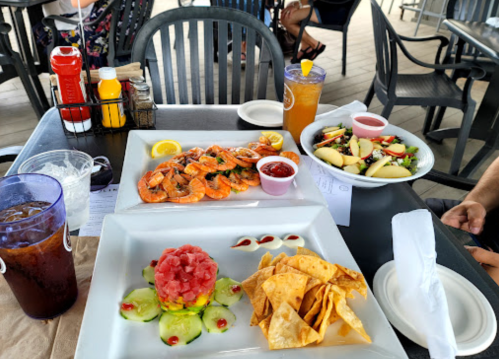 A table with shrimp, salsa, a salad, fruit, and tortilla chips, with drinks and condiments in a casual dining setting.