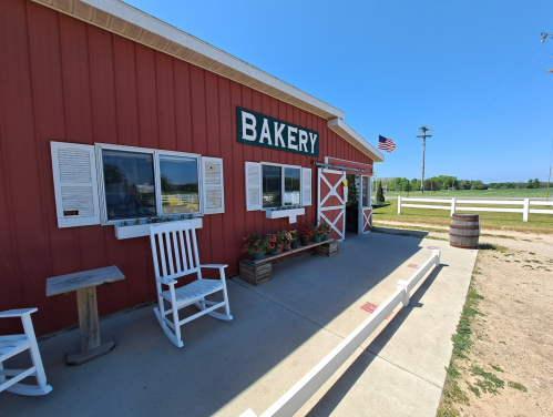 A red bakery building with white trim, rocking chairs, and flower boxes, set against a clear blue sky.