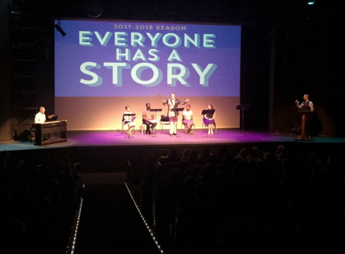 A stage performance with a large screen displaying "Everyone Has A Story," featuring musicians and performers.