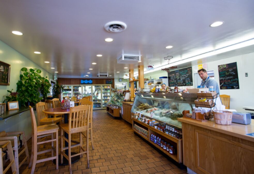 A cozy café interior with wooden tables, a display case of food, and a staff member serving customers.