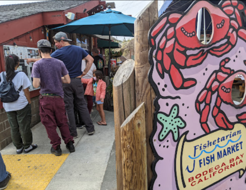 A colorful sign for a fish market in Bodega Bay, with people waiting in line outside under an umbrella.