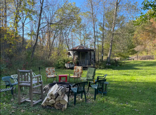 A cozy outdoor scene with a fire pit, chairs, and a small cabin surrounded by trees and autumn foliage.