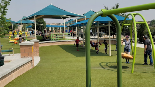 A playground with green swings, children playing, and shaded areas under blue canopies on a sunny day.