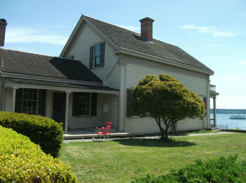 A historic house with a porch, surrounded by green grass and a tree, overlooking a body of water on a sunny day.