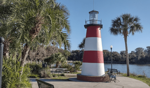 A red and white striped lighthouse surrounded by palm trees and benches near a calm waterway on a sunny day.