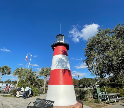 A red and white striped lighthouse stands against a blue sky, surrounded by palm trees and a park area.