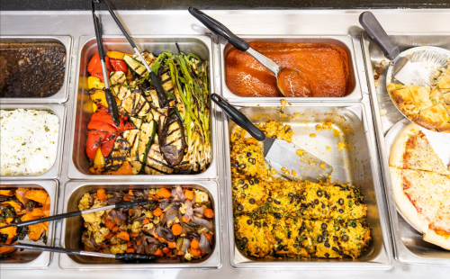 A buffet spread featuring grilled vegetables, rice, pasta, and pizza, with serving utensils in metal trays.