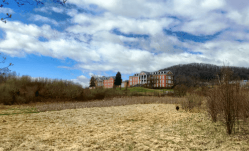 A large brick building sits on a hill, surrounded by fields and trees under a partly cloudy sky.