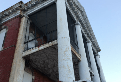 A weathered building with large columns, peeling paint, and vines growing on the lower balcony.