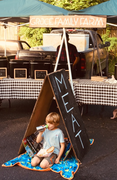 A child sits on a blanket under a sign reading "MEAT" at a farm stand, with a truck and food containers in the background.