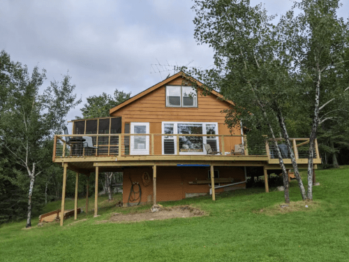 A two-story orange house on a raised deck, surrounded by green grass and trees under a cloudy sky.