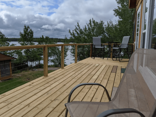 A wooden deck overlooking a lake, with two chairs and a small table, surrounded by trees under a cloudy sky.