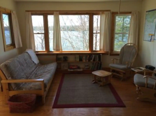 Cozy living room with a futon, rocking chair, and lake view through large windows. Warm wooden decor and a rug.