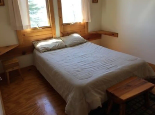 Cozy bedroom with a double bed, wooden accents, and natural light from two windows. Simple and inviting decor.