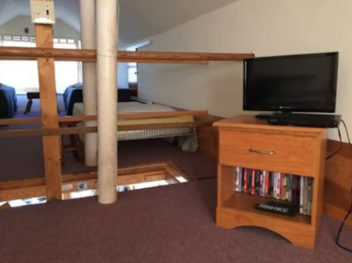 A cozy loft space with a wooden railing, a TV on a stand, and a view of two beds in the background.