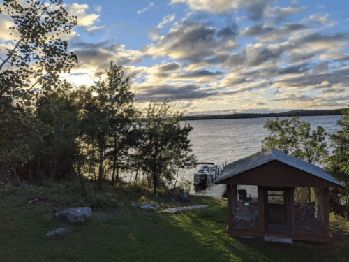A serene lakeside view at sunset, featuring a small cabin and a boat docked nearby, surrounded by trees and clouds.