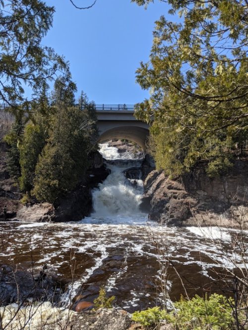 A waterfall cascades under a bridge, surrounded by lush greenery and a clear blue sky.