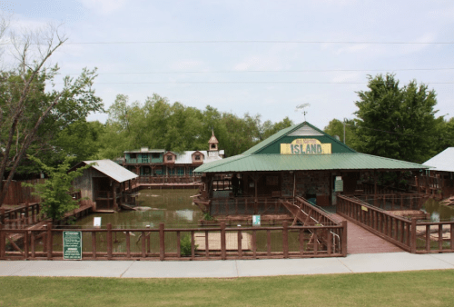 A wooden structure with a green roof overlooks a pond, surrounded by trees and buildings in a park-like setting.