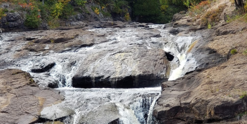 A cascading waterfall flows over rocky terrain, surrounded by lush greenery and rugged cliffs.