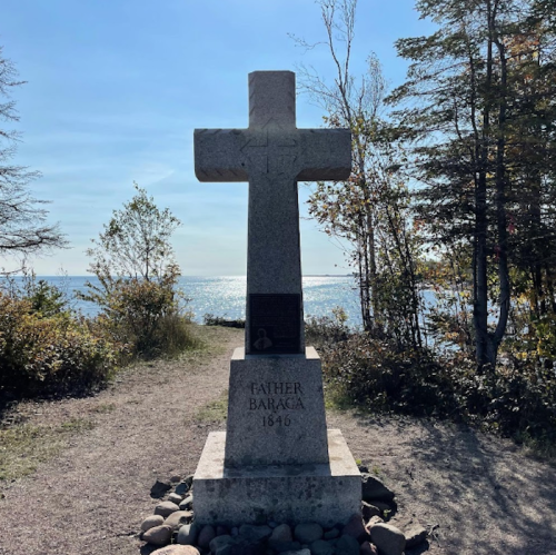 A stone cross monument stands by a path near the water, surrounded by trees under a clear blue sky.