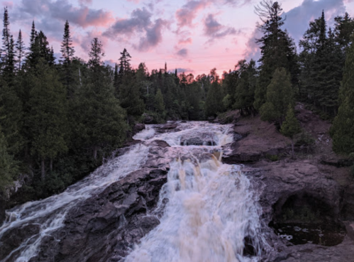 A waterfall cascades over rocky terrain, surrounded by lush trees under a colorful sunset sky.