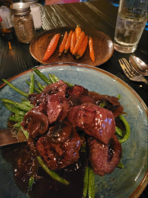 A plate of sliced beef in a dark sauce on green beans, with a side of glazed carrots in the background.