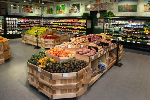A vibrant grocery store produce section with wooden displays of fruits and vegetables, including bananas, tomatoes, and sweet potatoes.