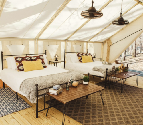 Cozy glamping tent interior with two beds, wooden tables, and warm lighting in a forest setting.