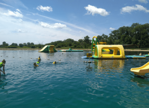 Children in life jackets play on inflatable water structures in a sunny lake setting.