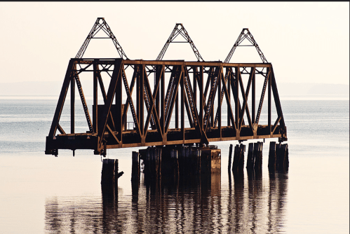 Rusty train trestle bridge stands over calm water, reflecting the structure against a soft, hazy background.