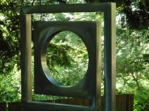 A large metallic sculpture featuring a square frame with a circular cutout, surrounded by lush greenery.