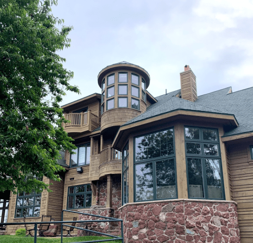 A large wooden house with a stone base and multiple windows, featuring a round tower and surrounded by trees.