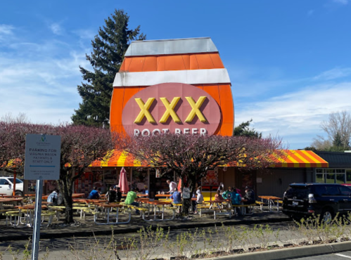 A colorful building with "XXX Root Beer" signage, outdoor seating, and trees in bloom on a sunny day.