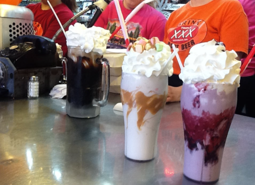 Three colorful milkshakes topped with whipped cream and garnishes, served at a counter with staff in bright shirts.