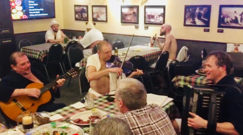 A lively restaurant scene with musicians playing instruments while diners enjoy their meals at a checkered tablecloth setting.