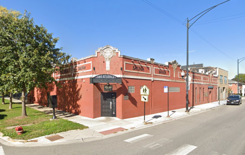 A red brick building with "Sauna" and "Sweat Lodge" signs, located at a street corner with trees nearby.