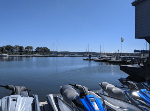 A serene marina scene with jet skis in the foreground and boats docked under a clear blue sky.