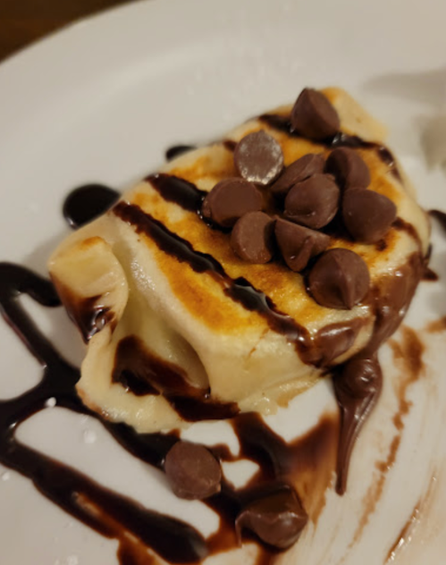 A dessert featuring a golden-brown pastry topped with chocolate chips and drizzled with chocolate sauce on a white plate.